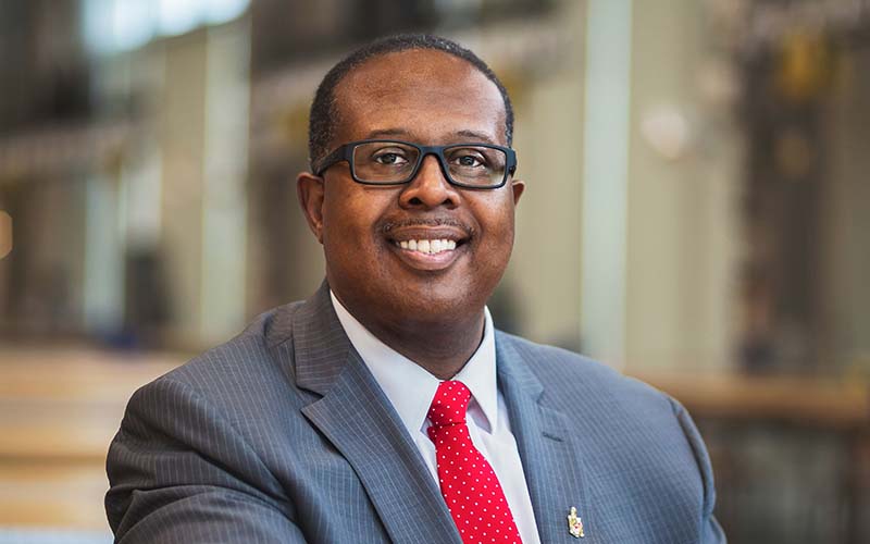 Dean Samuel Graham, Jr. is smiling. He is wearing a grey suit, a white shirt, and a red tie.