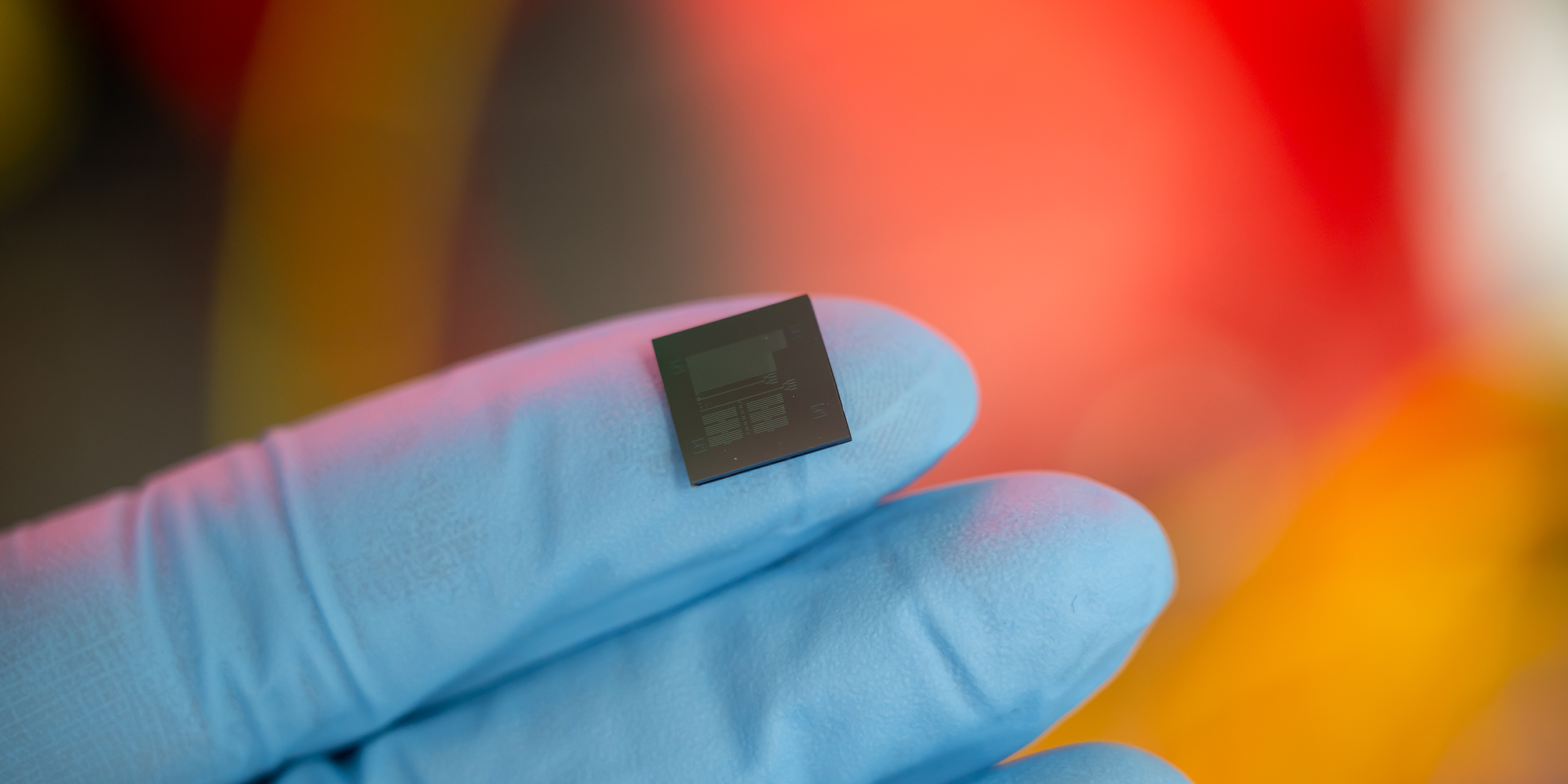 two fingers wearing blue latex gloves holding a small semiconductor chip in front of an obscured orange and red background