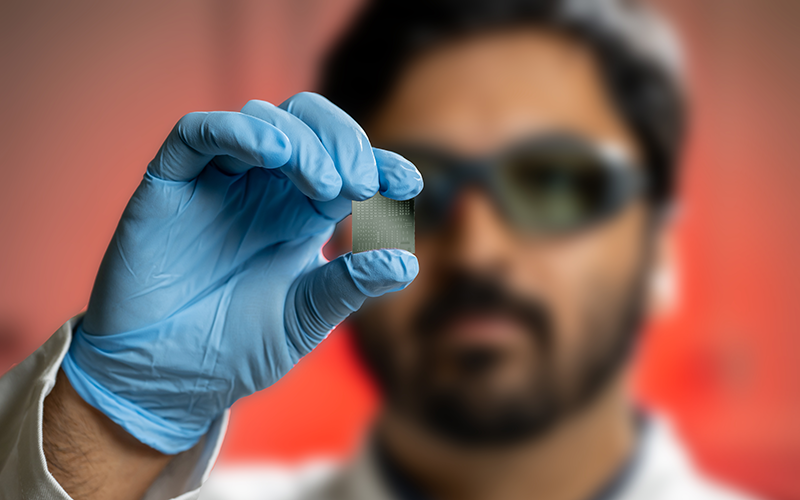 a Maryland engineer holds a thin, silver semiconductor chip in front of his face between his index finger and thumb with a blue glove on