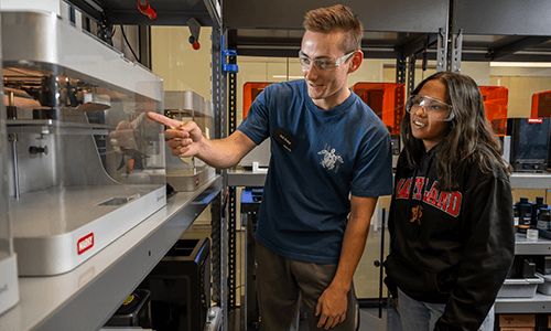 two students observe a metallic colored 3-D printer at the TerrapinWorks lab